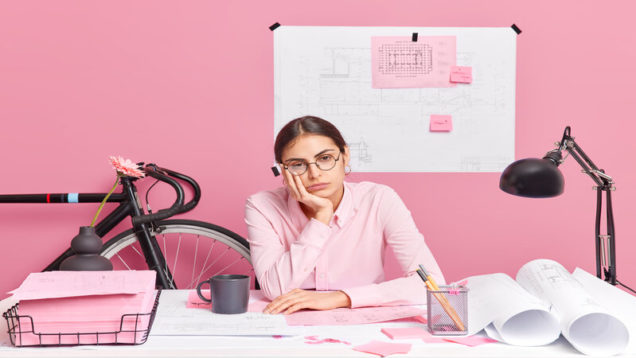 Sad tired woman wears spectacles poses at desktop works all day long on blueprints involved in learning process isolated over pink background prepares building project feels exhausted during deadline